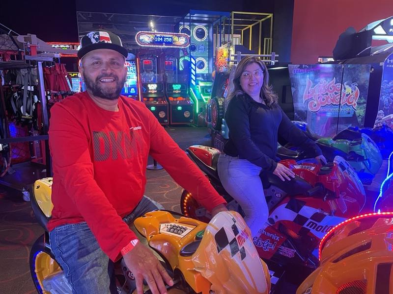 Two people on arcade motorcycle games, smiling.