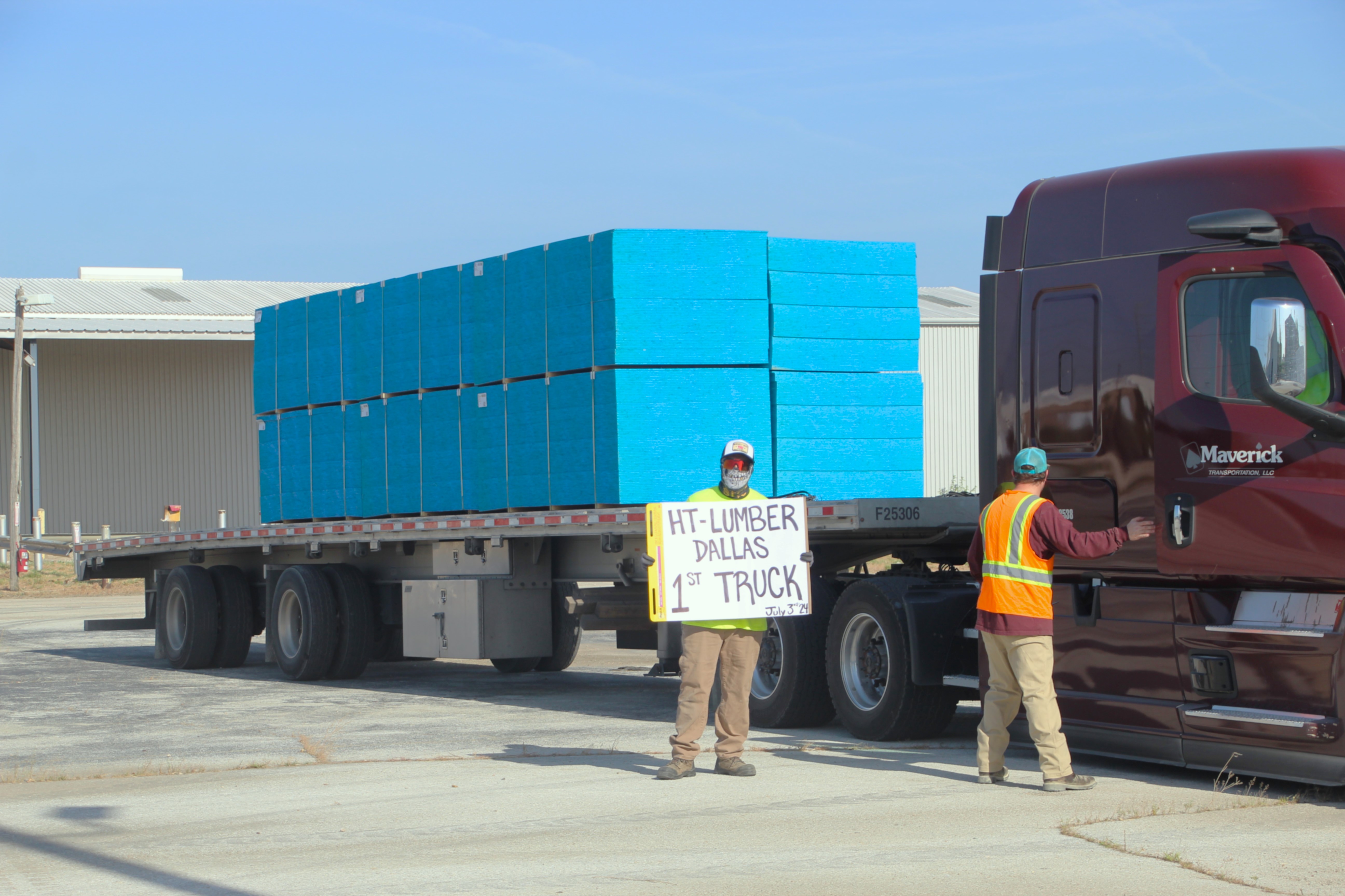 Worker holds 'HT Lumber Dallas 1st Truck' sign by a loaded truck.