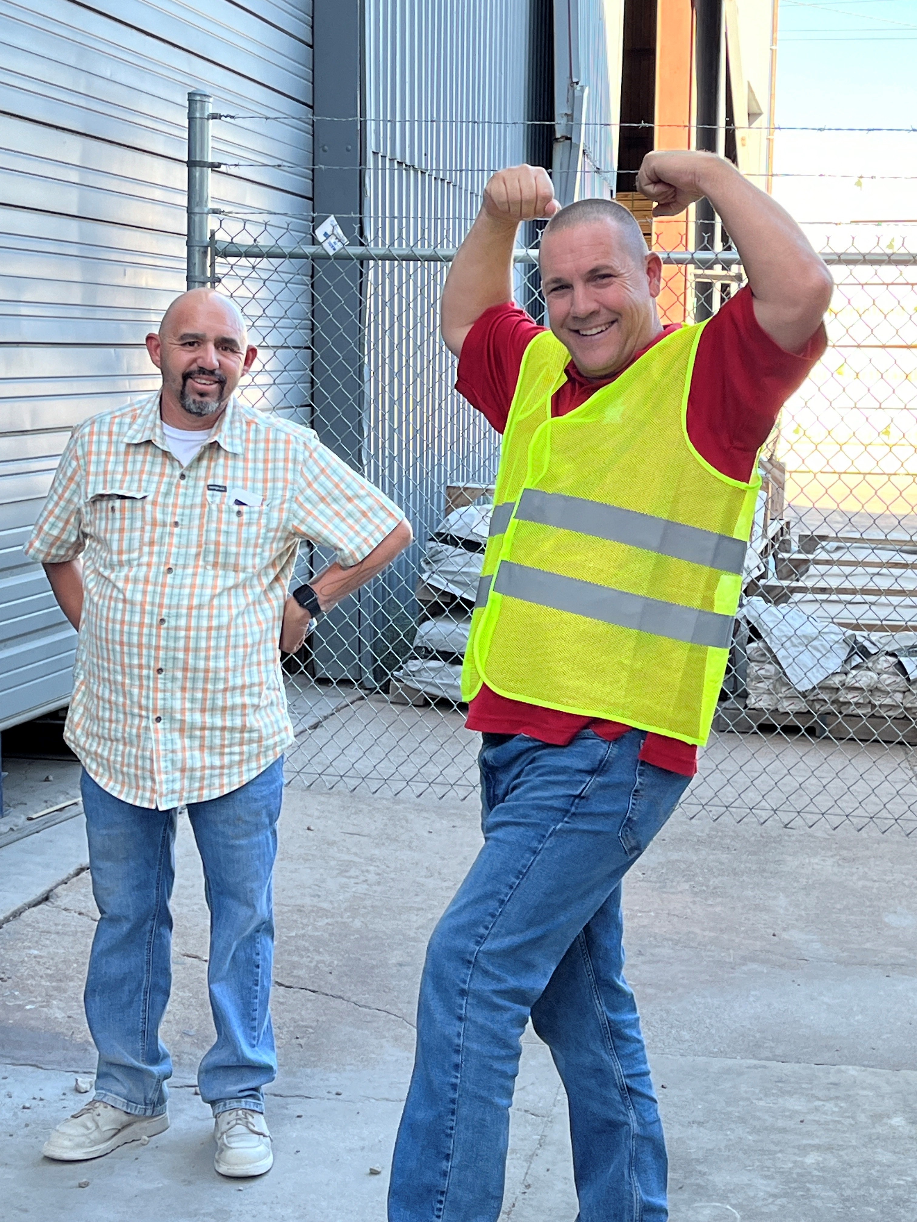 Man flexing playfully next to another by a fence.